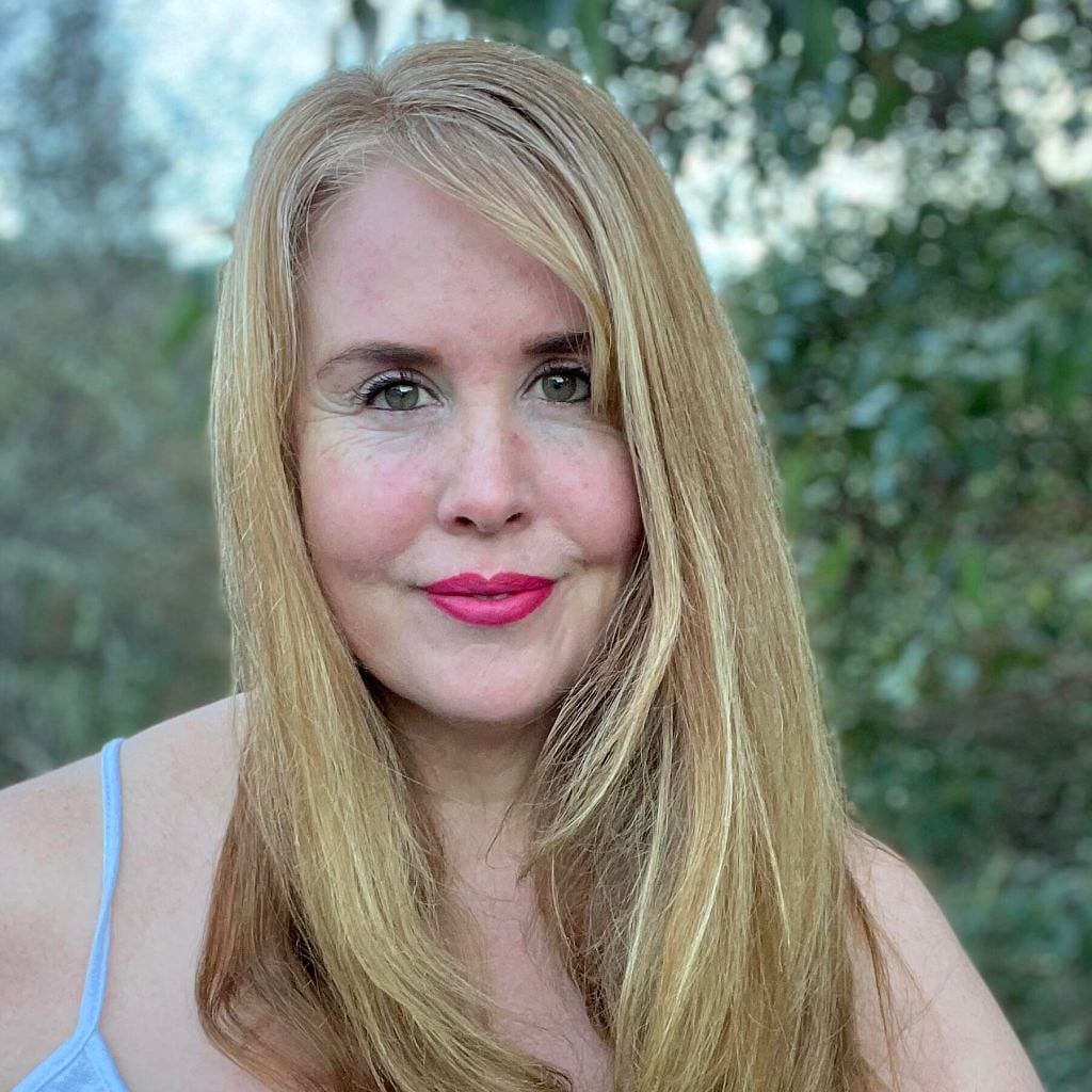 A woman with long blonde hair smiling outdoors in front of greenery