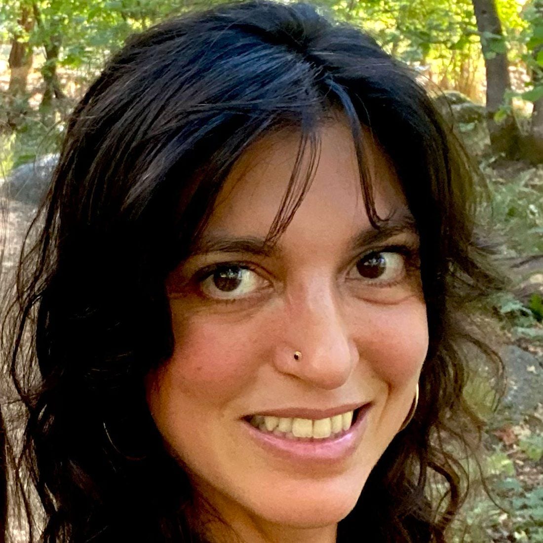 A smiling woman with dark wavy hair outdoors among trees